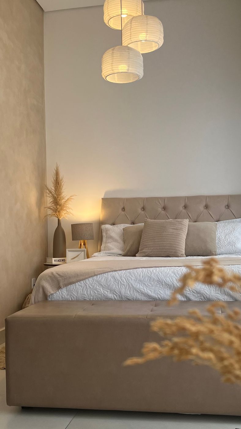 Cenário cama, com cabeceira capitonê, roupa de cama clara, luminárias e vaso com pampas