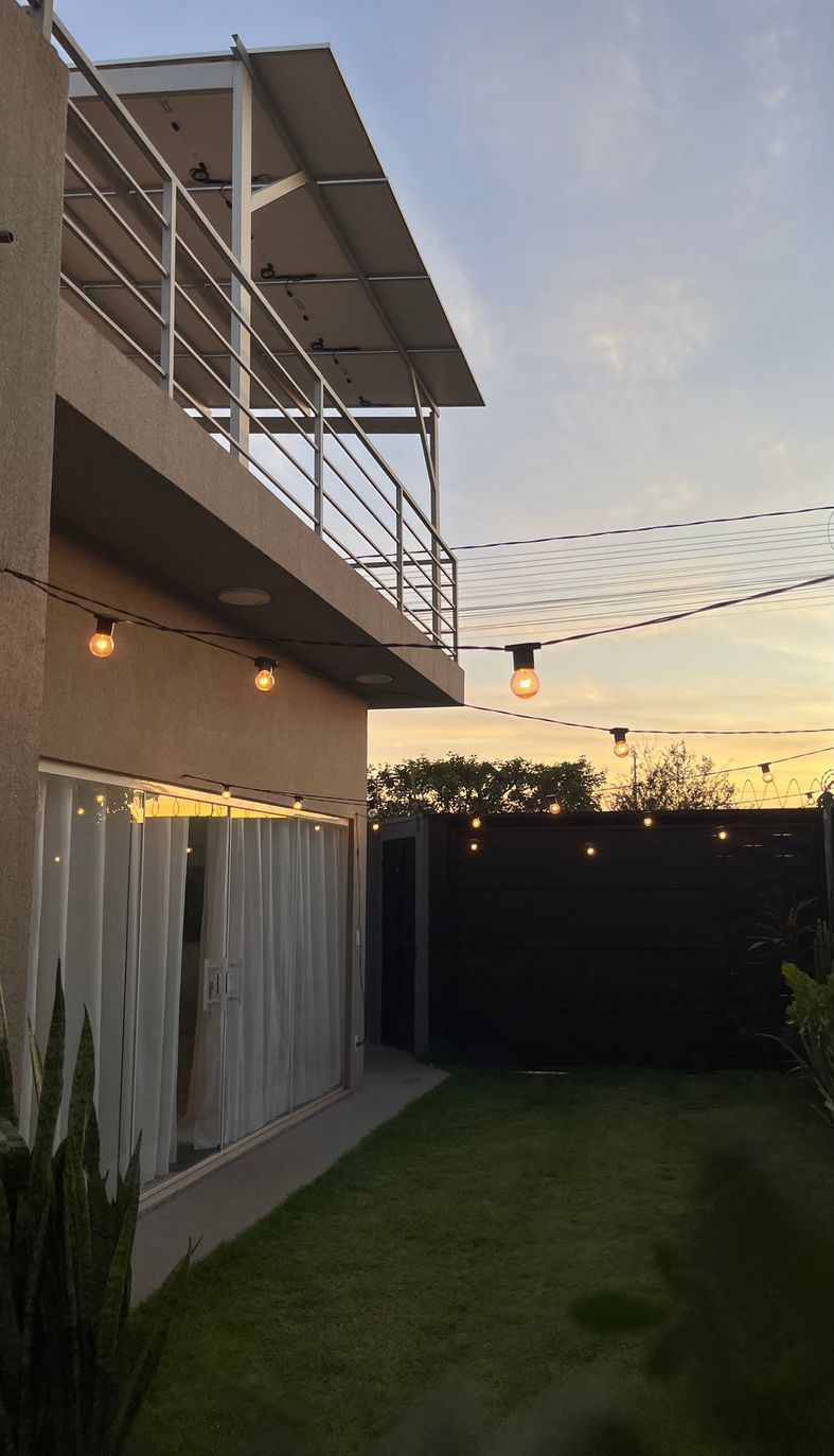 Área externa do estúdio ao entardecer, com gramado, terraço no segundo piso e varal de luzes acesas